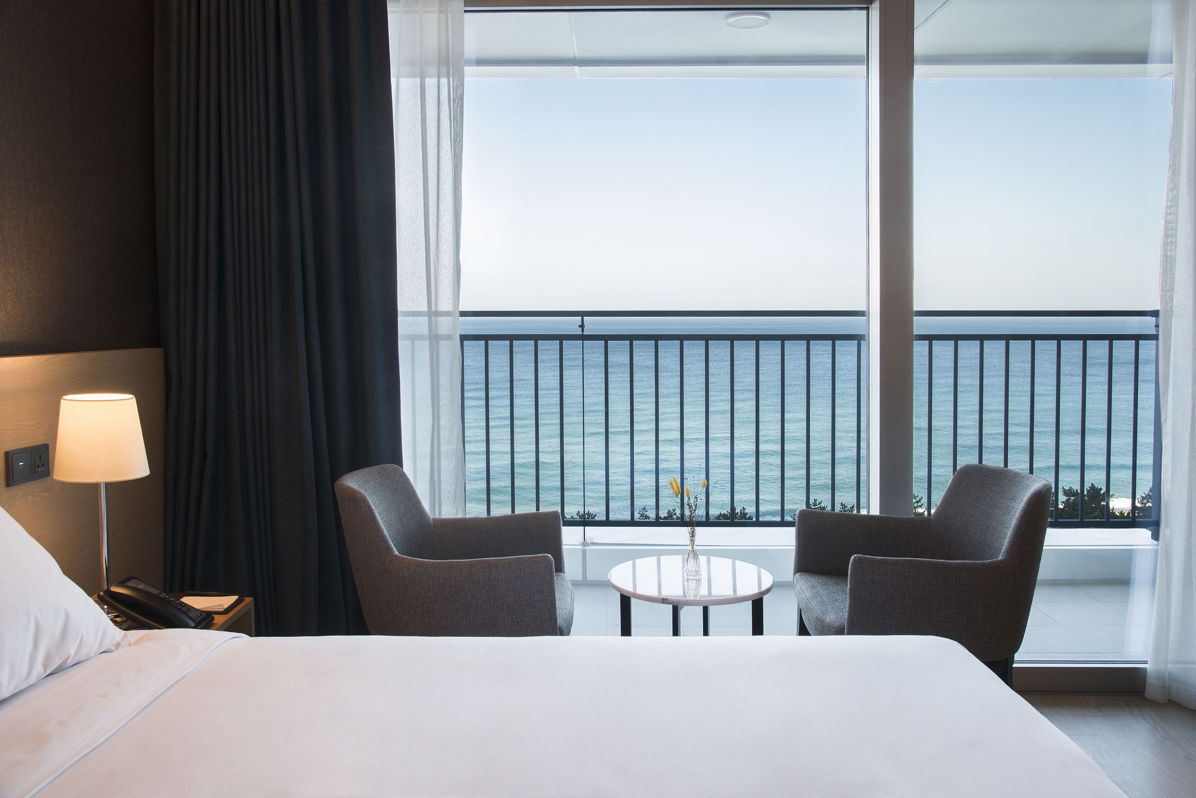 A hotel room with a bed, two armchairs, a small table, and a view of the sea through large windows.