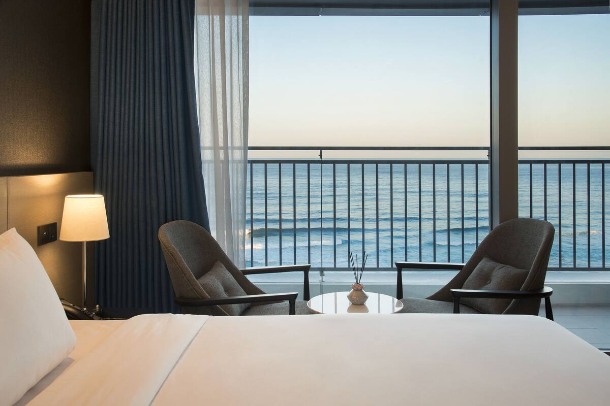 A hotel room with a bed, two armchairs, a small table, and a view of the ocean through a balcony railing.
