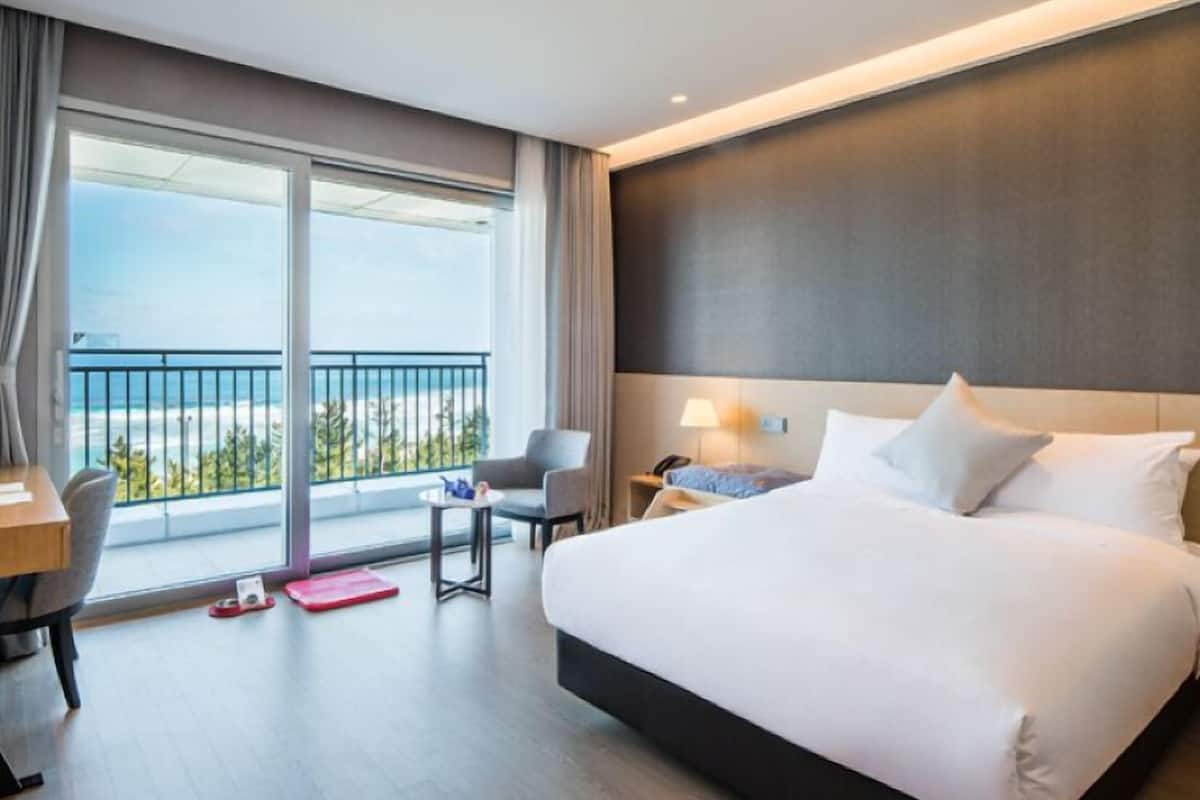 A hotel room with a large bed, a desk, a chair, a small table, and a view of the beach through a balcony.