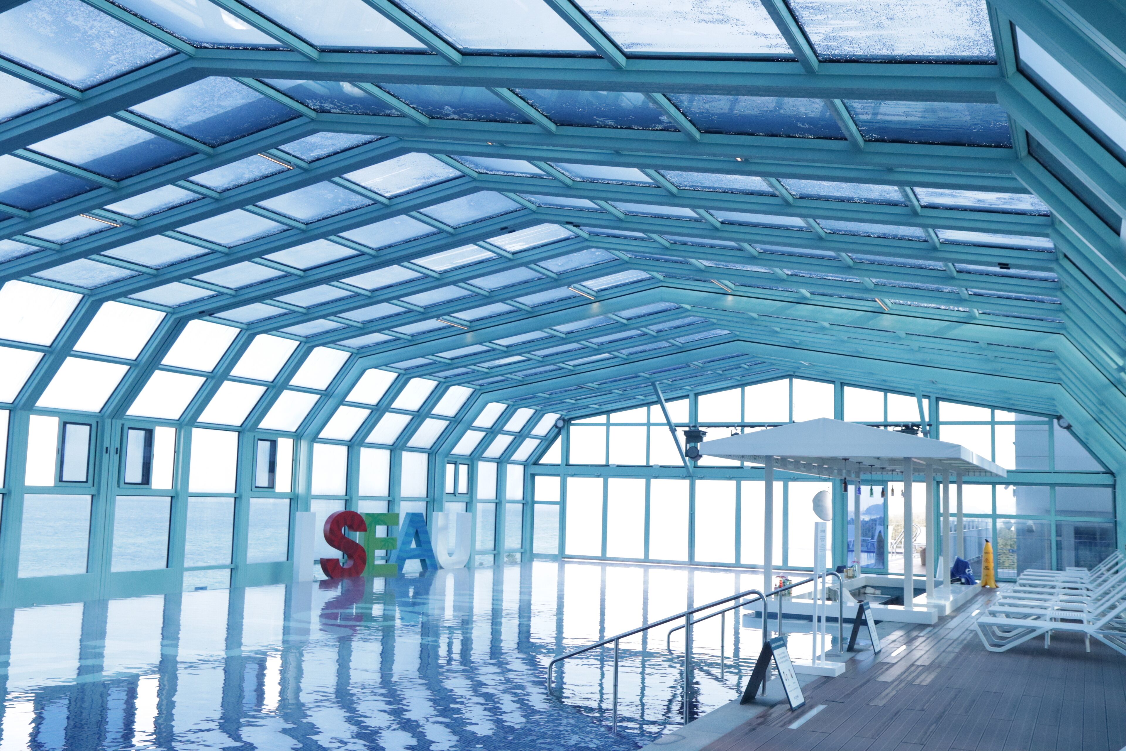 An indoor swimming pool with a glass ceiling, a large 'SEA' sign, and lounge chairs along the poolside.