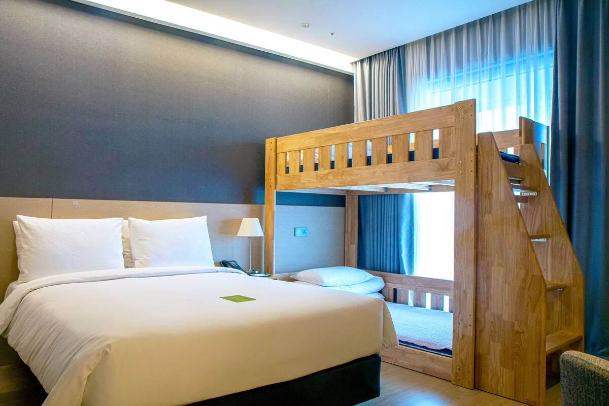 A hotel room with a bunk bed, a large bed, a bedside table with a lamp, and a view of the outdoors through a window with curtains.