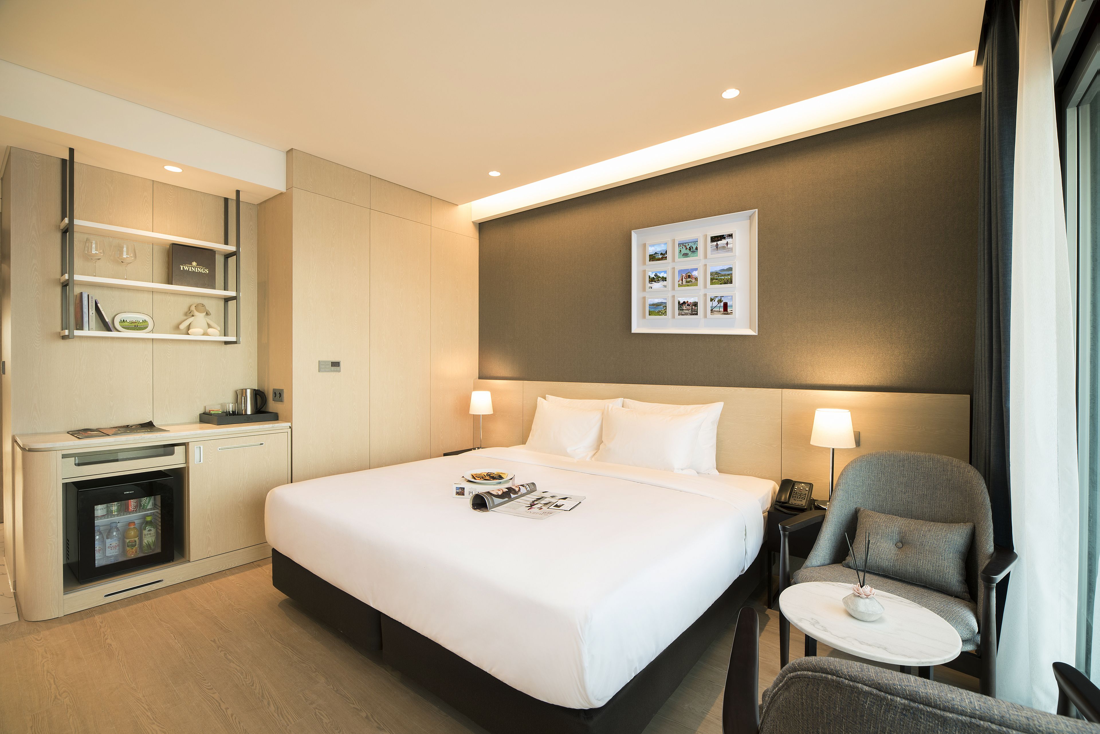 Habitación de hotel con cama, mesita de noche, silla, minibar y un expositor de fotos en la pared.