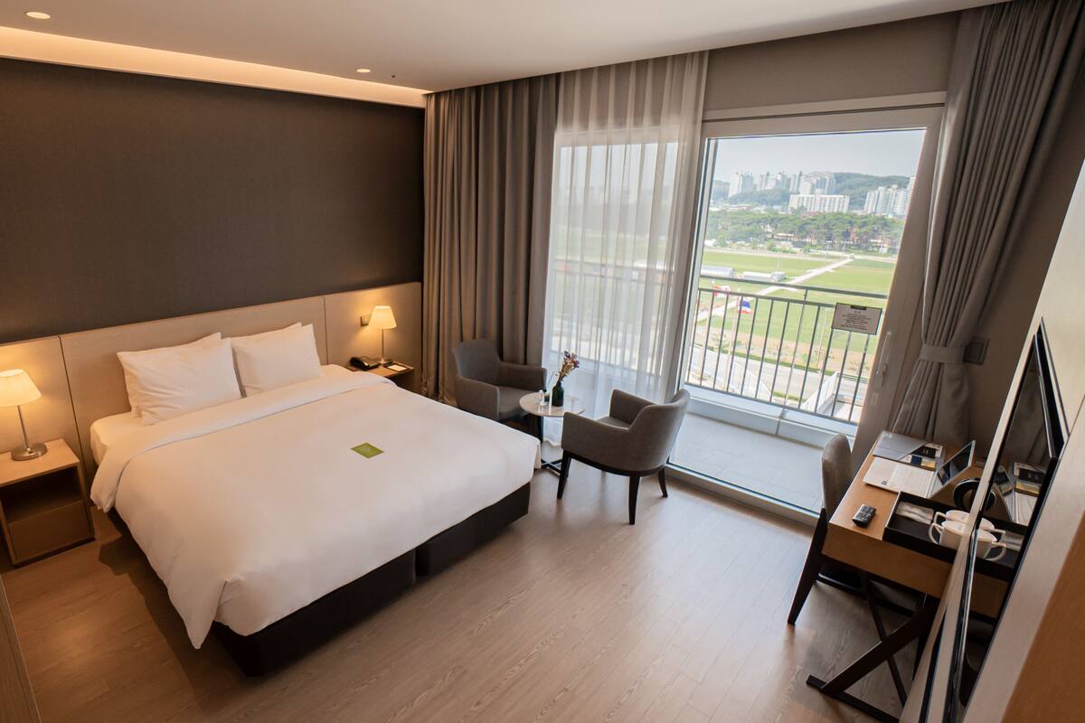 A modern hotel room with a large bed, a desk, and a chair. There is a view of a cityscape through the window.