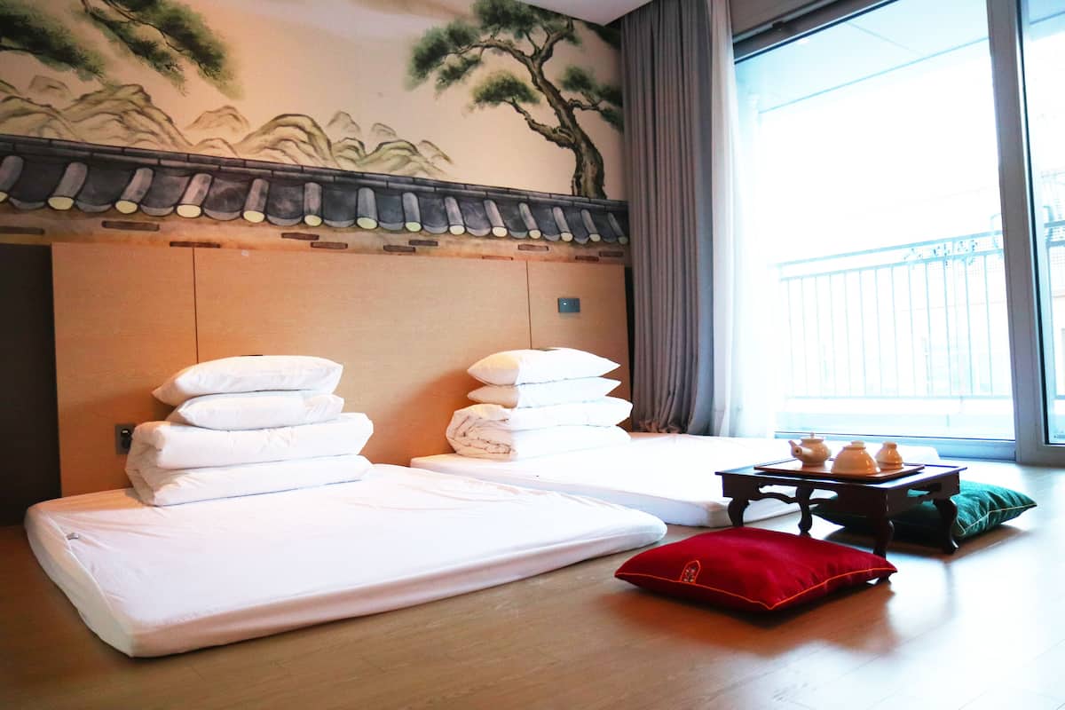 A hotel room with two beds, a small table with teapots, and a view of the outside balcony.