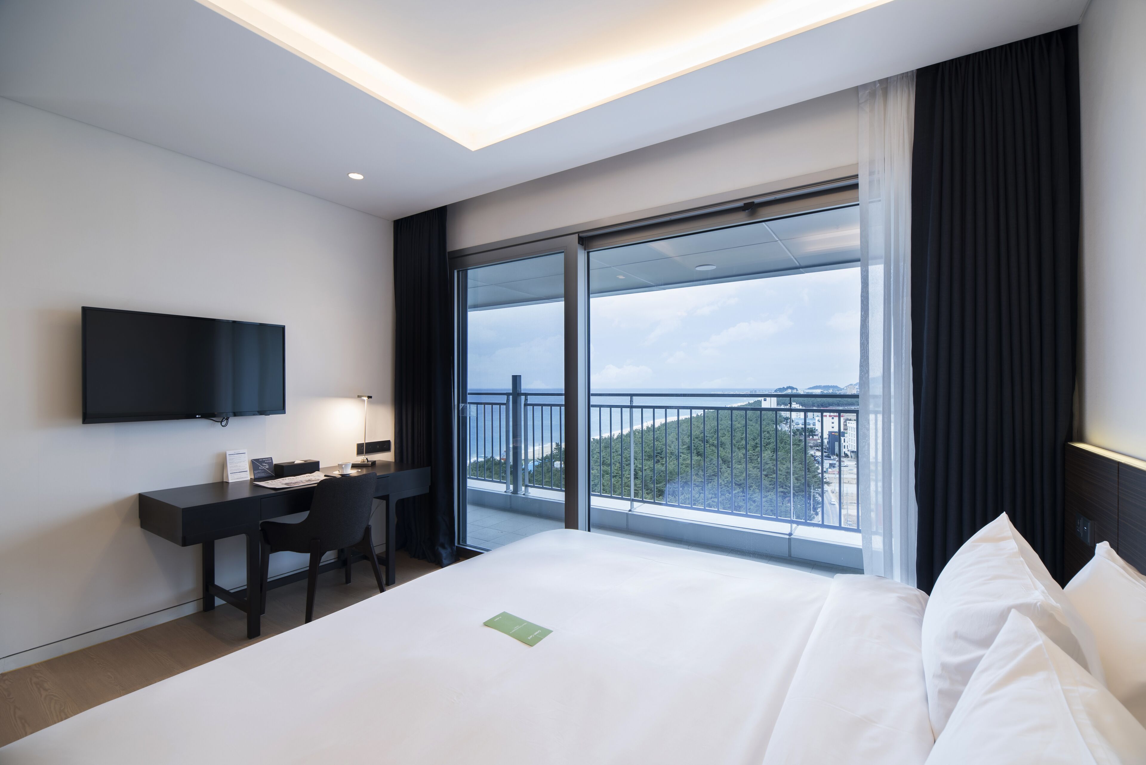 A modern hotel room with a large bed, a desk with a chair, a flat-screen TV, and a balcony with a city view.