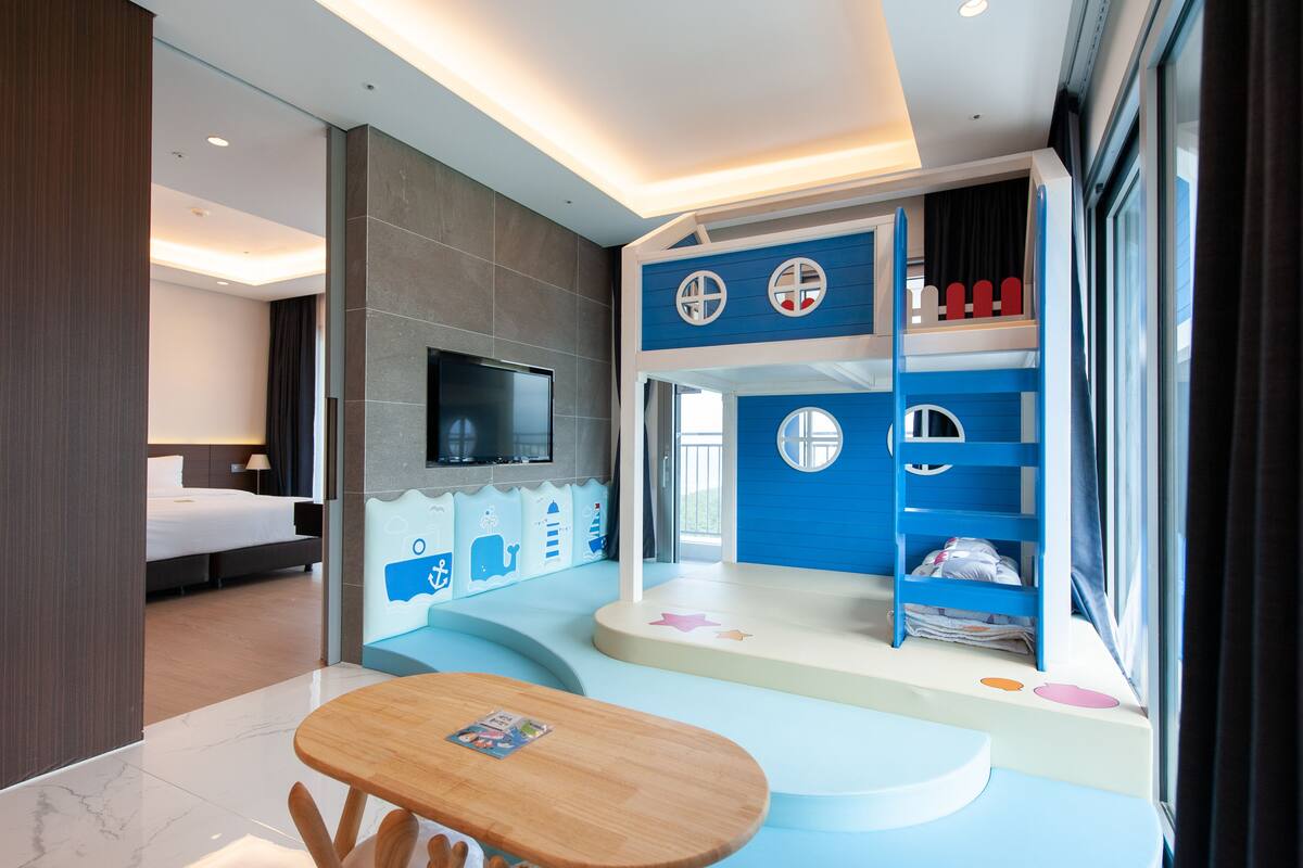 A modern hotel room with a large bed, a TV, a children's play area, and a dining table.
