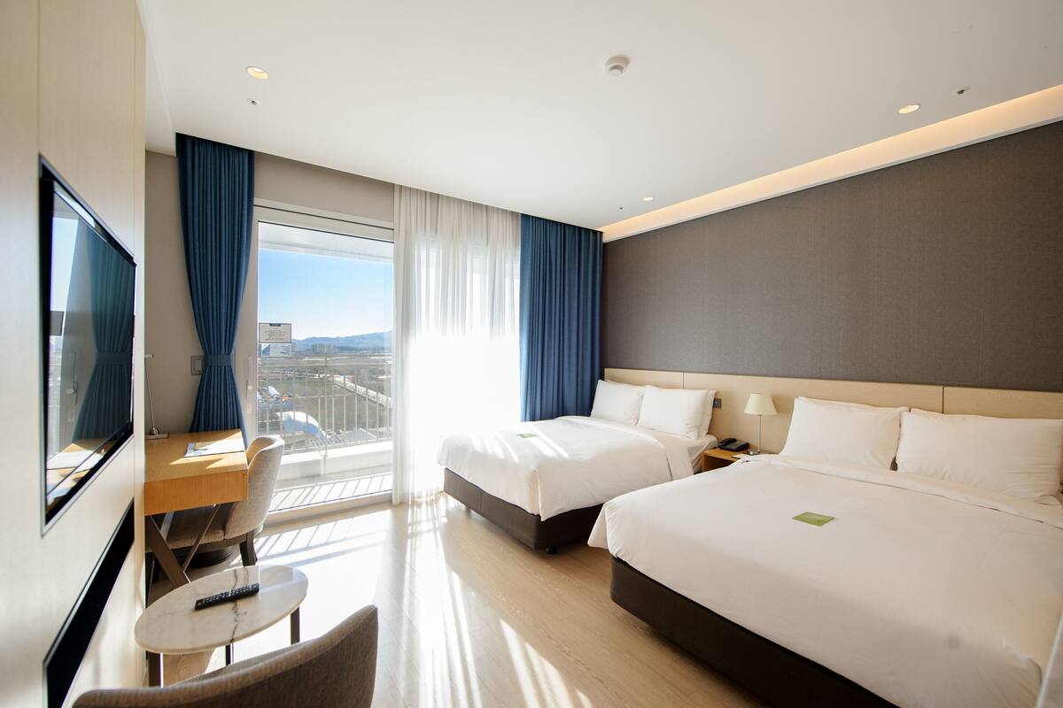 A hotel room with two beds, a desk, a chair, a TV, and a large window with curtains.