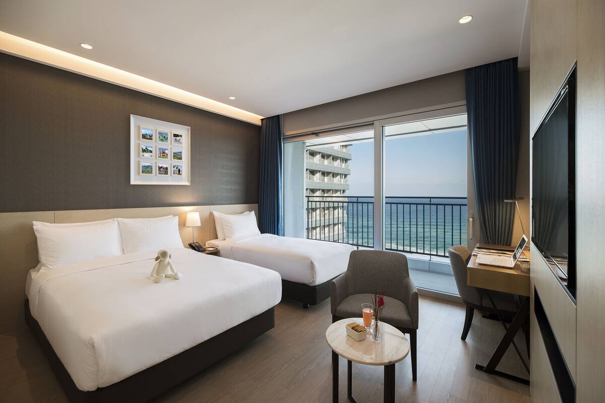 A hotel room with two beds, a desk, a chair, a small table, and a large window with a view of the sea.