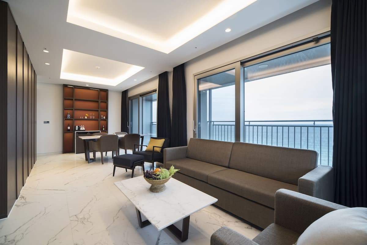 A modern living room with a sofa, dining table, and chairs. There is a view of the sea through large windows.