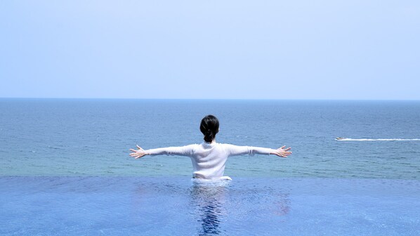 A person standing at the edge of an infinity pool with arms outstretched, overlooking a calm sea.