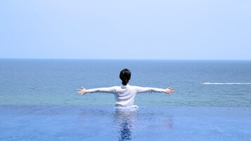 A person standing at the edge of an infinity pool with arms outstretched, overlooking a calm sea.