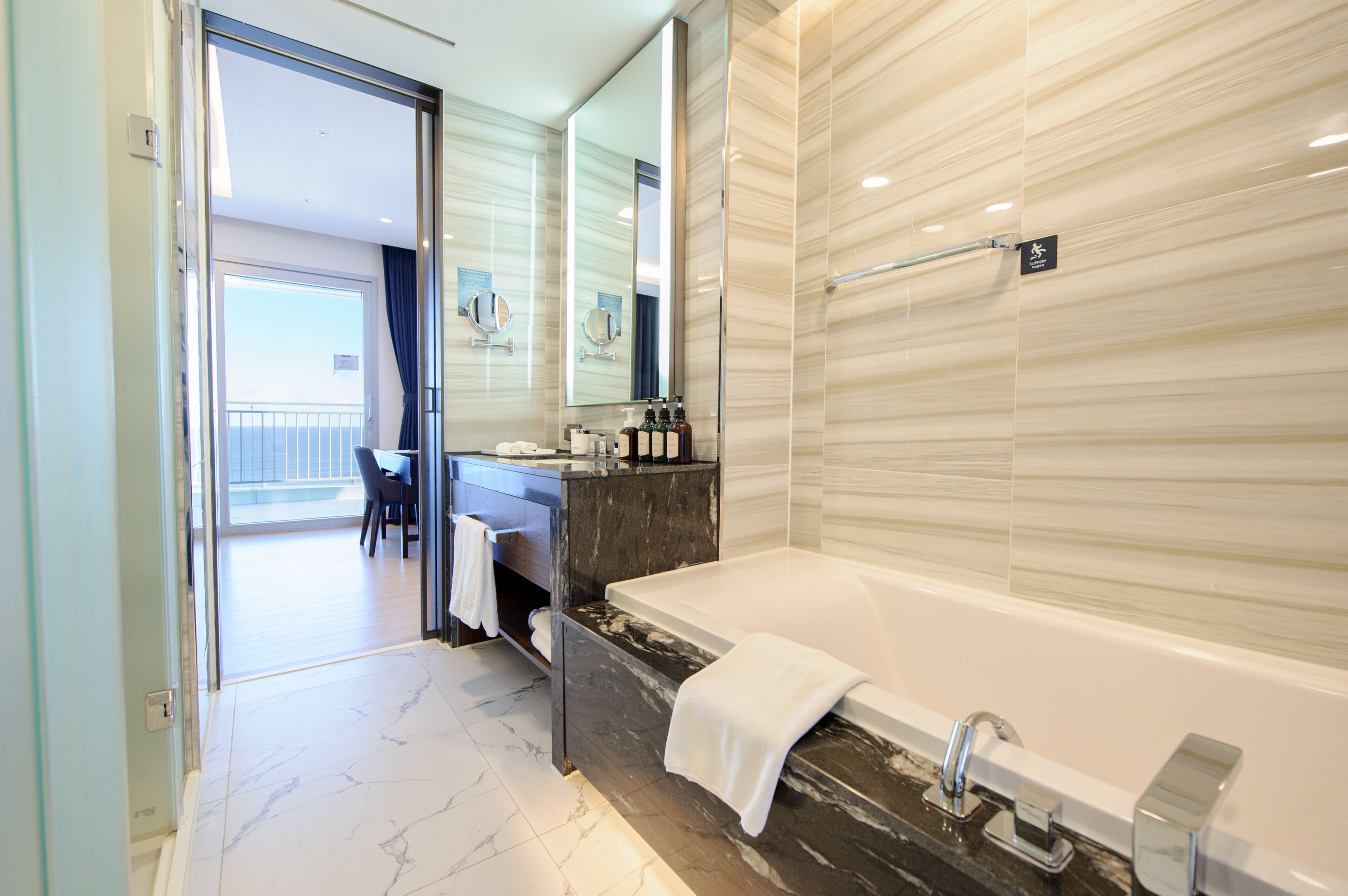 Ein modernes Badezimmer mit Marmorboden, einer großen Badewanne, einem Waschtisch mit Spiegel und Blick auf einen Balkon.