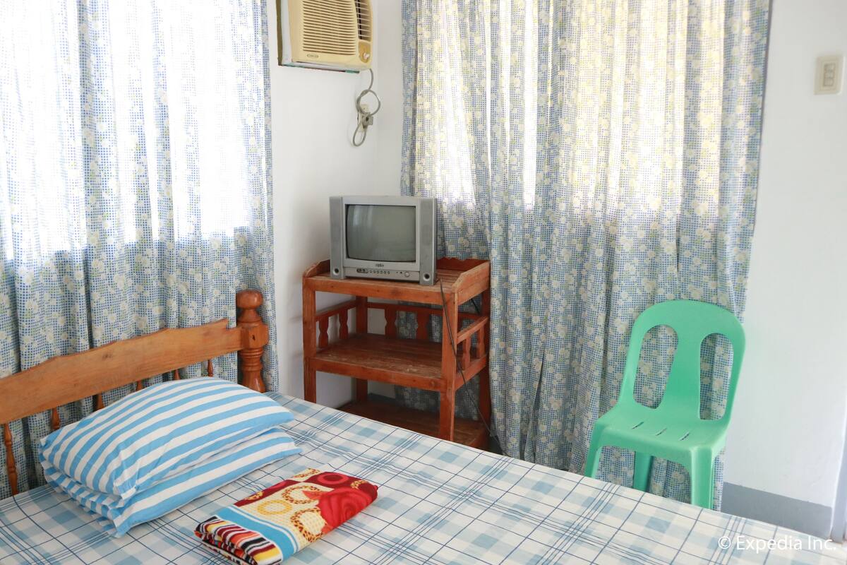Un dormitorio con una cama, un televisor sobre un soporte de madera, una silla verde y cortinas.