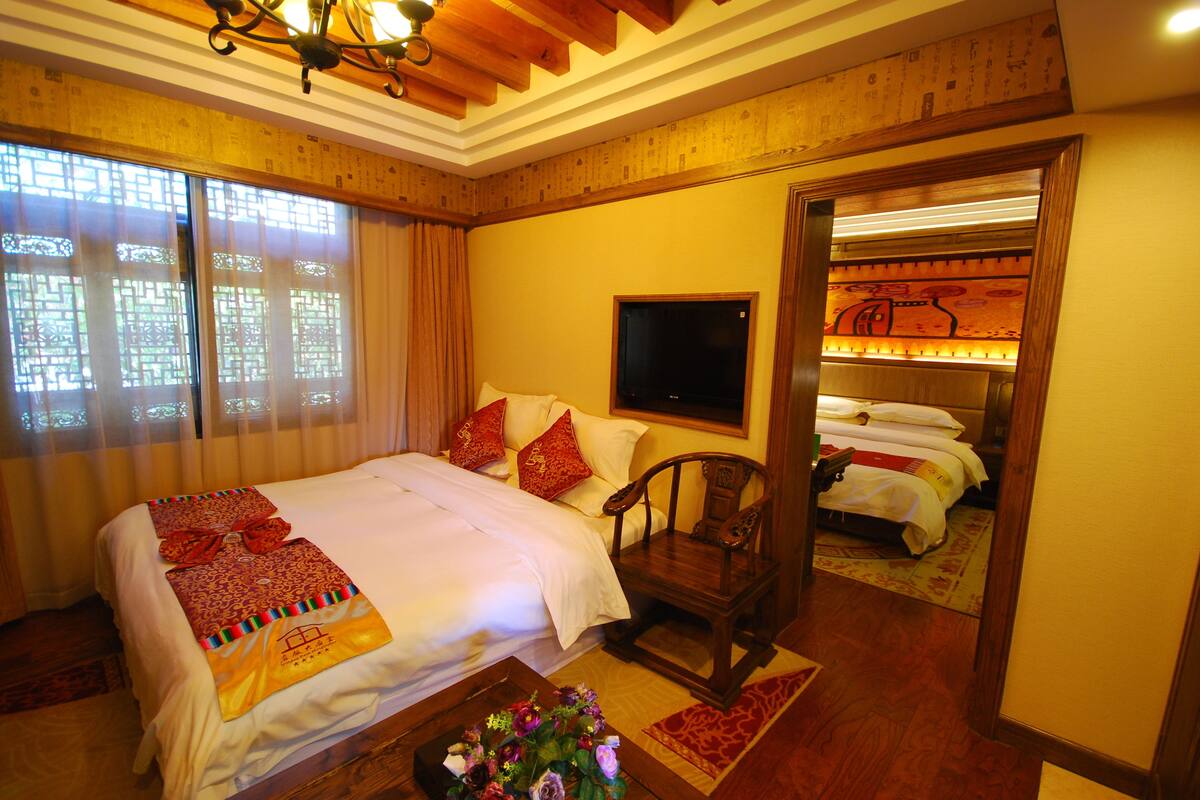Quarto de hotel com uma cama grande, uma cadeira de madeira, uma televisão e uma mesinha com flores.