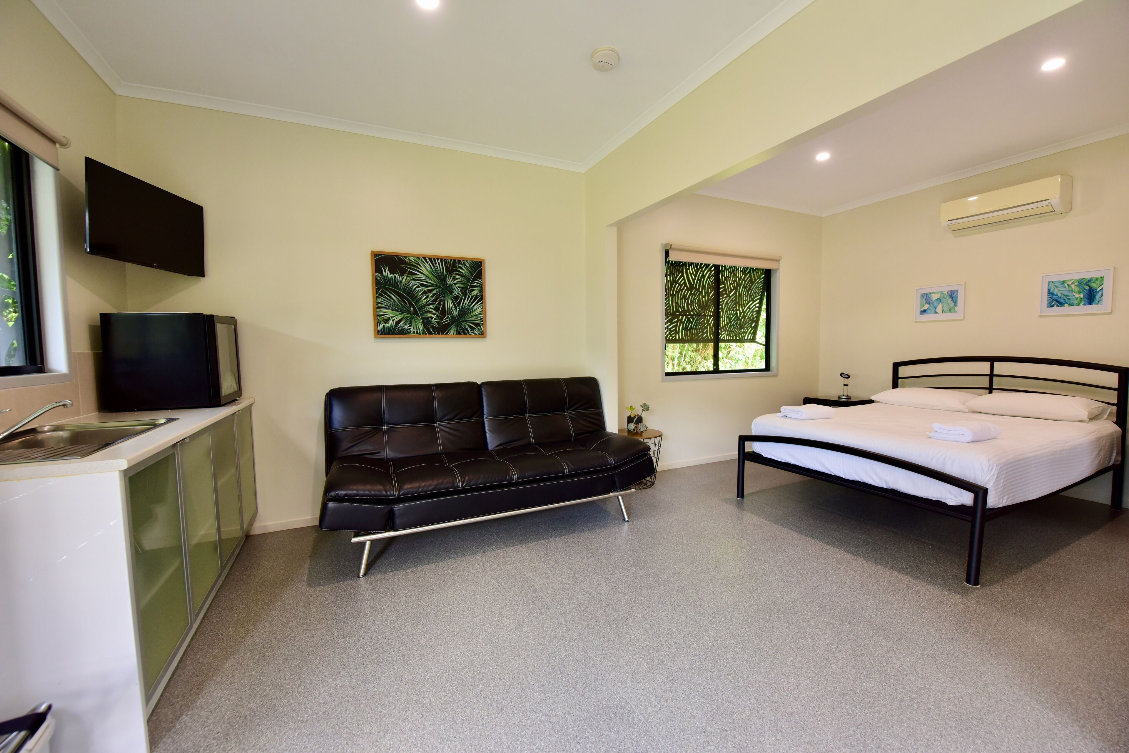 Une chambre moderne avec un canapé en cuir noir, un lit avec du linge de maison blanc, un téléviseur à écran plat fixé au mur et une fenêtre donnant sur un paysage verdoyant.