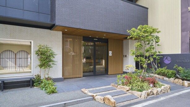 A hotel entrance with a modern design, featuring a wooden facade, a glass door, and a landscaped garden area with rocks and plants. - Hotel ASIATO Namba (Osaka)