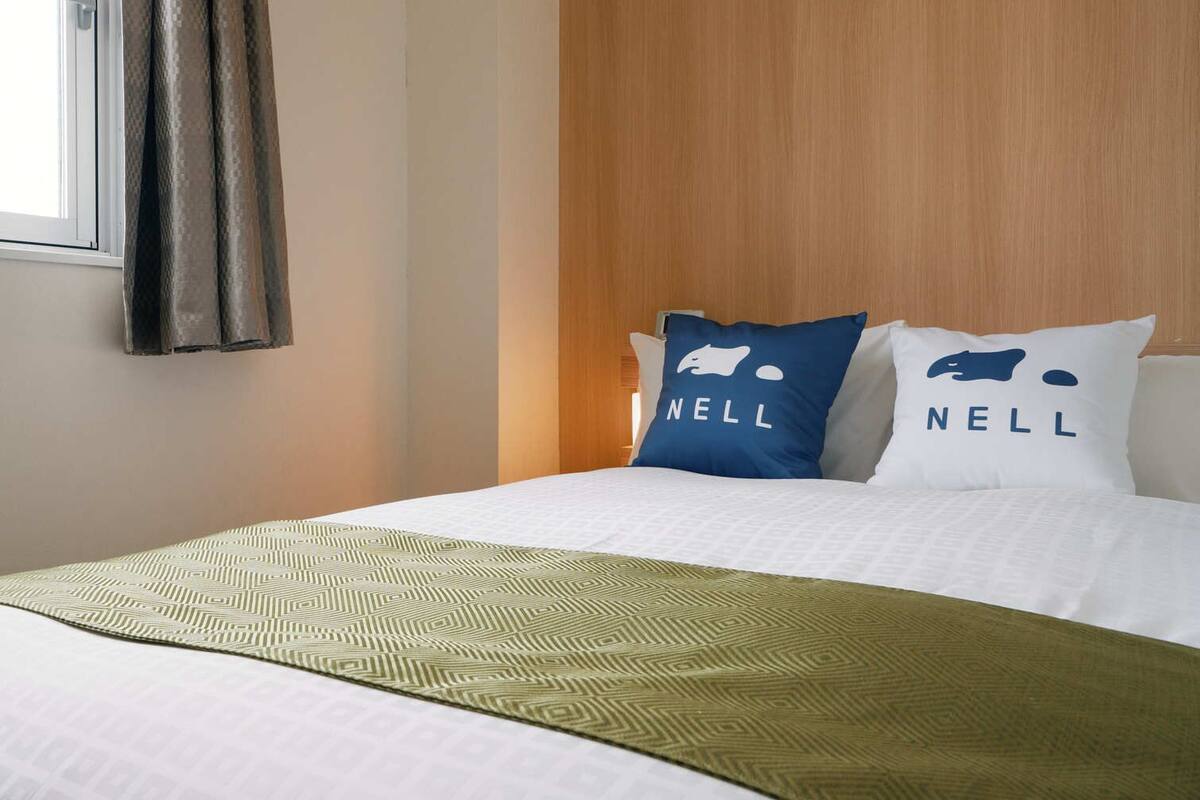 A hotel room with a bed, two pillows with the brand name 'NELL,' a window with curtains, and a patterned throw blanket.