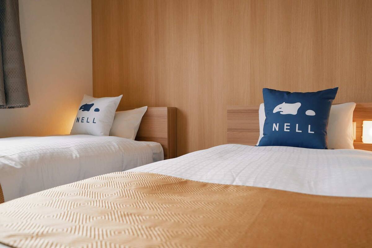 A hotel room with two beds, each with a pillow featuring the word 'NELL' and a logo. The room has wooden headboards and a large bed with a patterned bedspread.