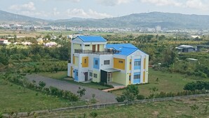 Um edifício de vários andares com telhado azul, rodeado por vegetação e uma paisagem montanhosa.