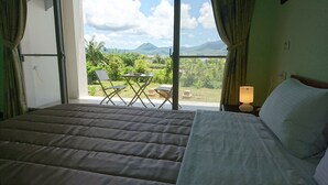 Quarto de hotel com cama, uma mesinha com cadeiras e vista para vegetação e montanhas através de grandes janelas.