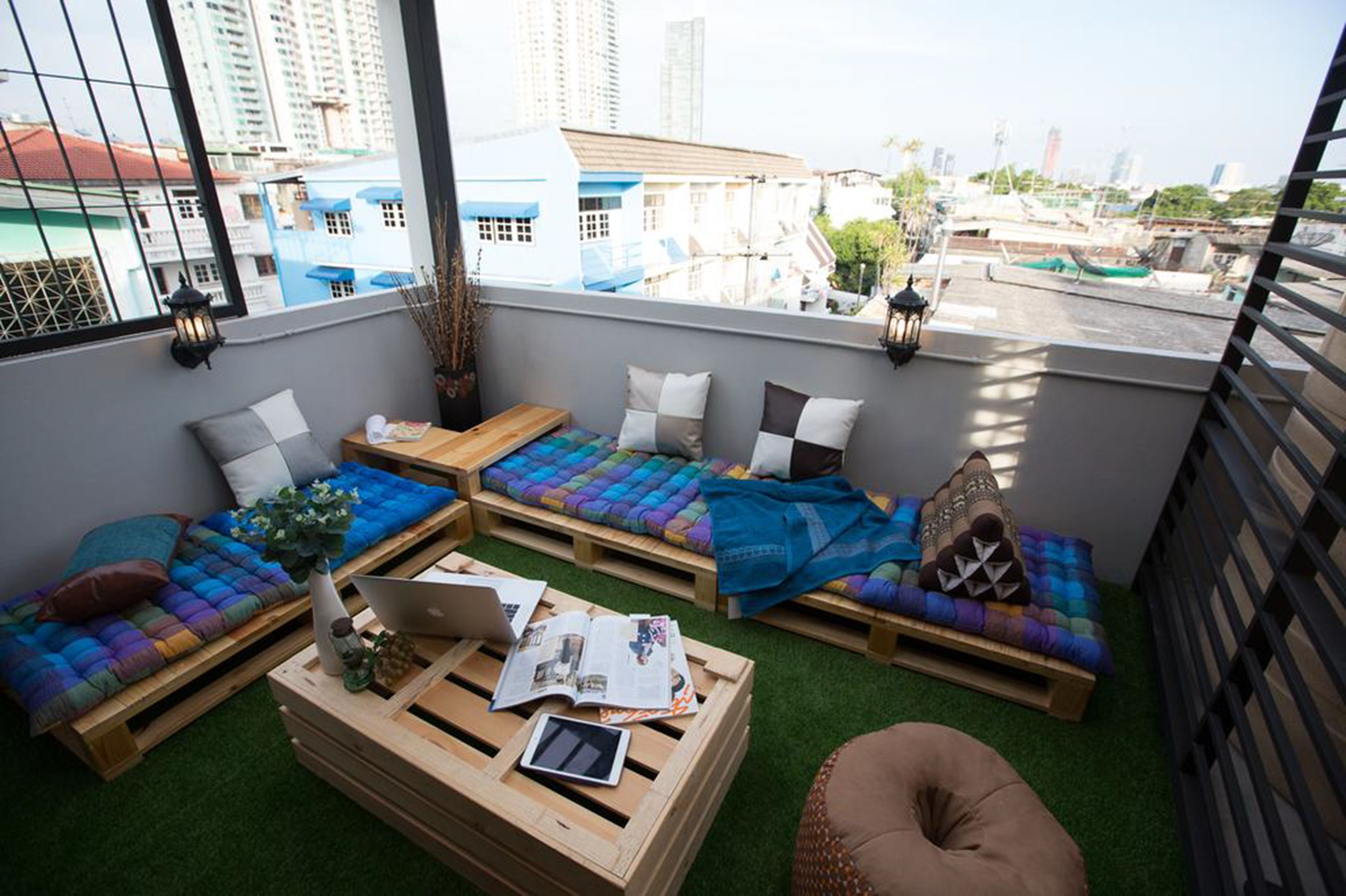 A cozy balcony with a view of the city, featuring a sofa set, a small table with a laptop and magazines, and a decorative lantern.
