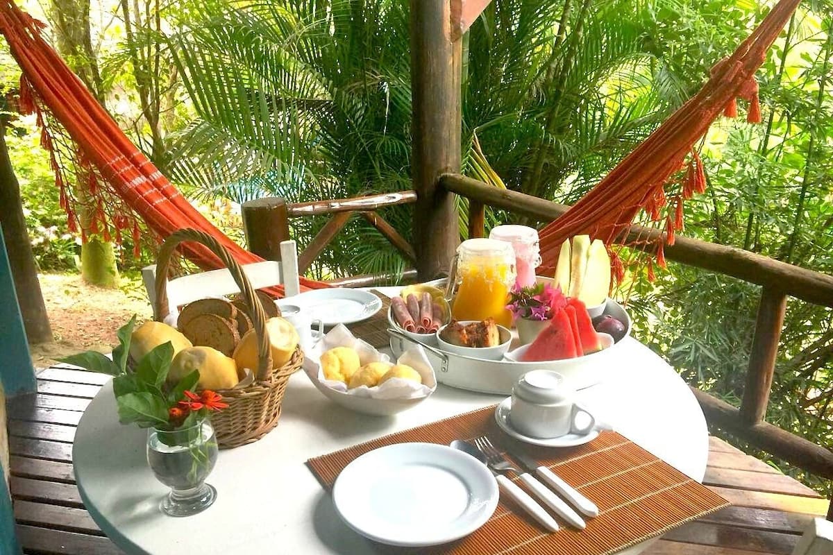 A table set outdoors with breakfast items including bread, fruit, and a drink, surrounded by lush greenery.