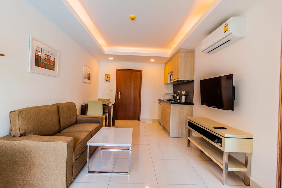 A modern living room with a sofa, coffee table, and TV. There is a kitchen area with cabinets and a microwave.