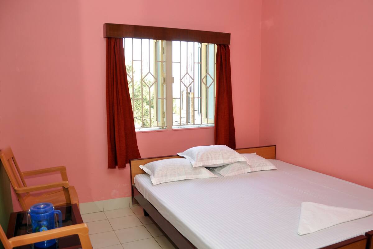 A bedroom with a single bed, a chair, a table with a blue water bottle, and a window with red curtains.