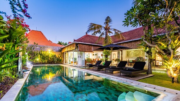 A swimming pool with lounge chairs and umbrellas, surrounded by lush greenery and a building with a tiled roof. - Villa Livo (Denpasar)