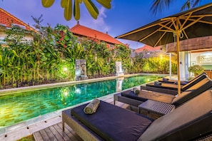 A swimming pool with lounge chairs and an umbrella, surrounded by lush greenery and a building with a red roof.