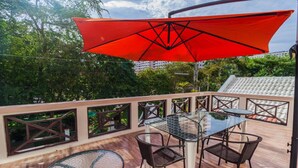 Une terrasse extérieure avec une table et des chaises en verre, un parasol rouge et une vue sur la verdure et les bâtiments.