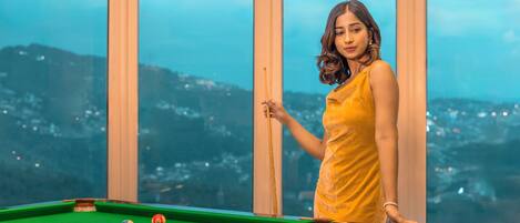 Una persona con un vestido amarillo de pie junto a una mesa de billar y vista panorámica de la ciudad.