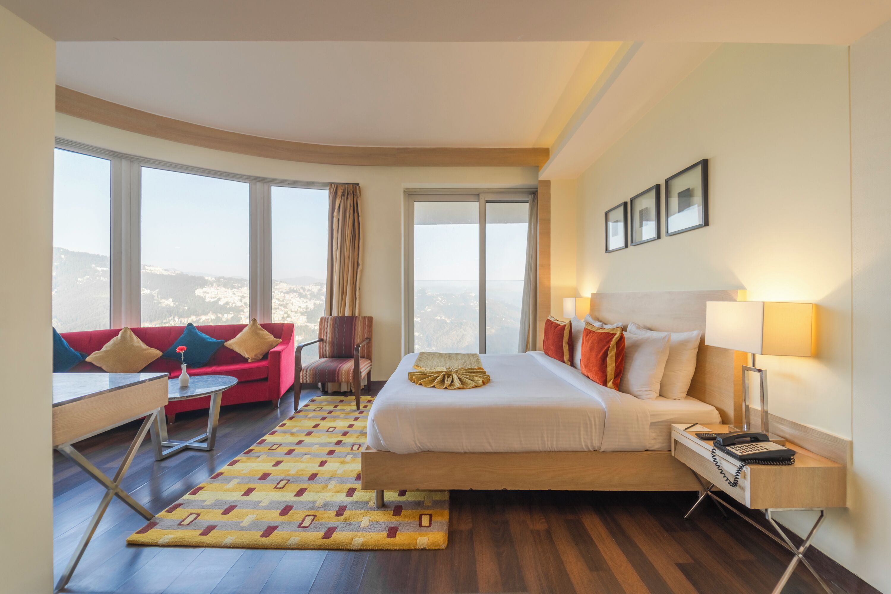 A hotel room with a large bed, a red sofa, a small table, and a view of the city through large windows.