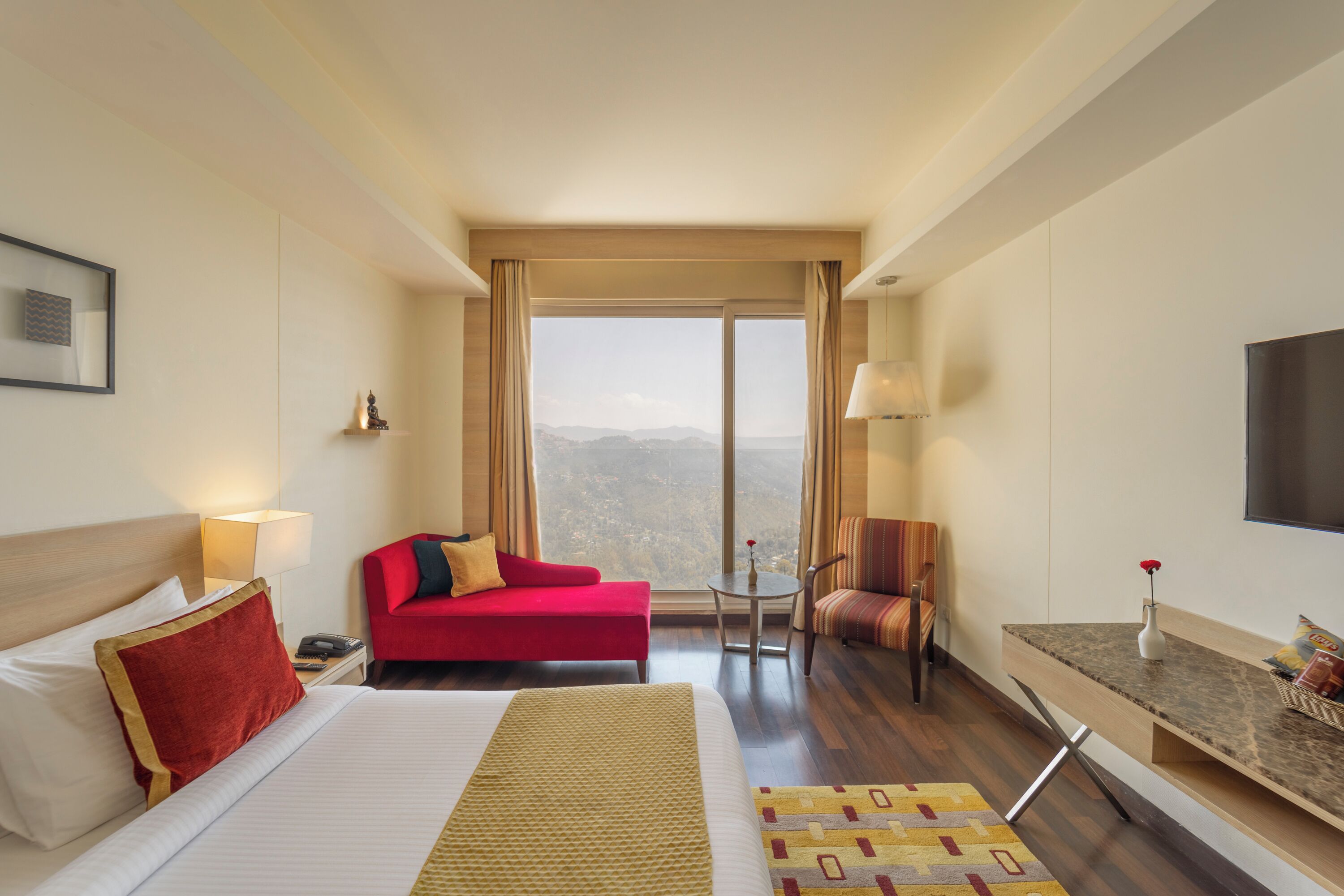 A hotel room with a large bed, a red sofa, a small table with a lamp, a TV, and a view of the mountains.