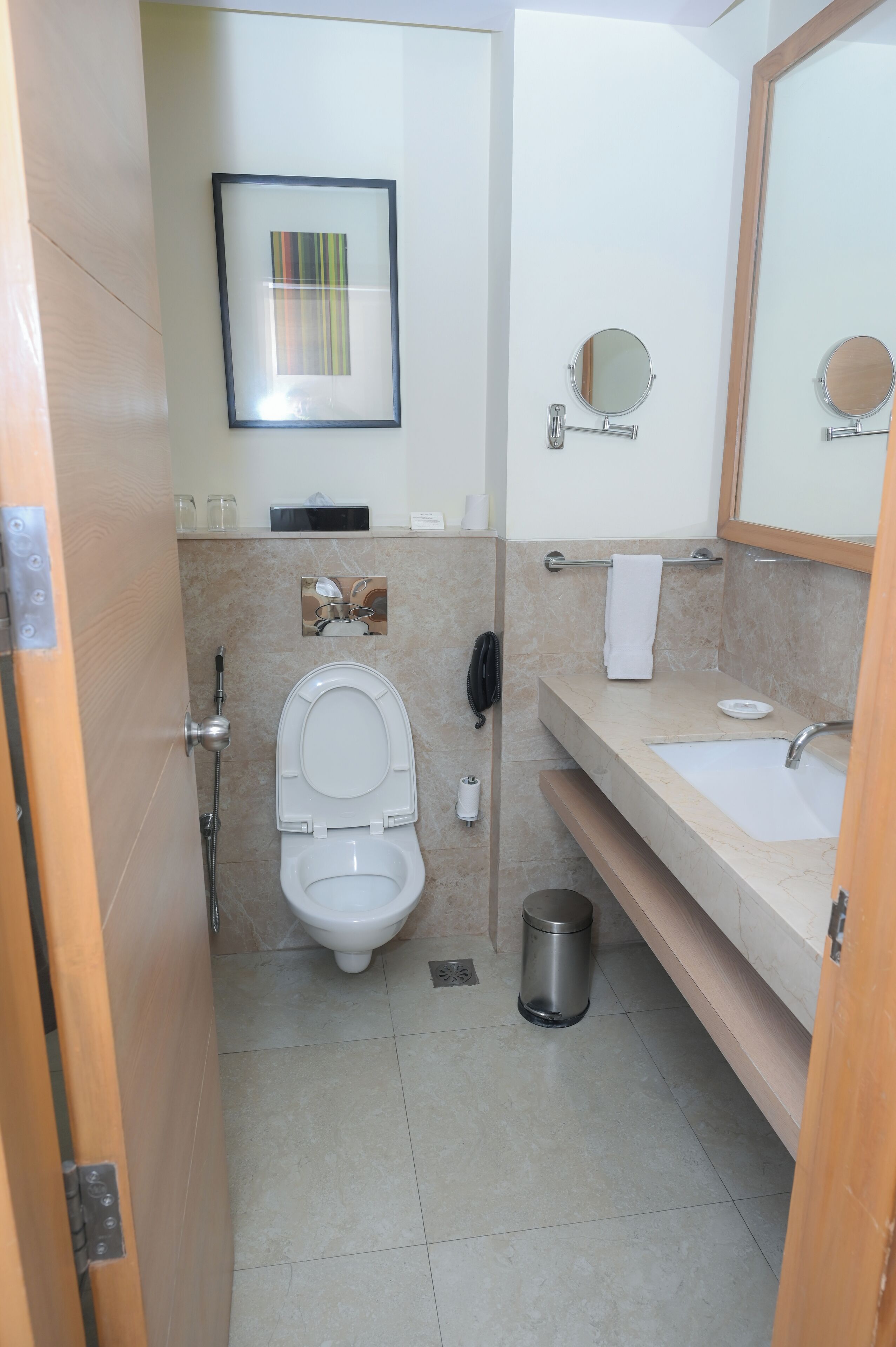 A bathroom with a toilet, sink, and mirror. There is a trash can and a towel rack.
