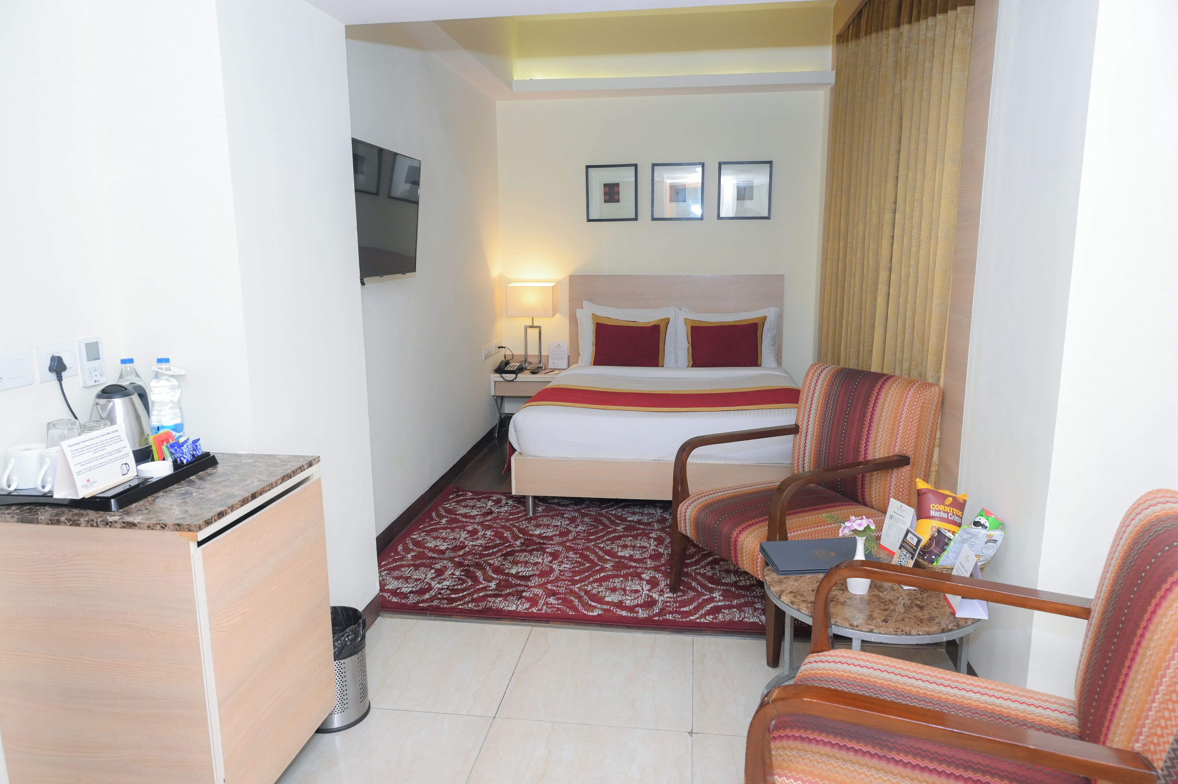 A hotel room with a bed, a desk with a telephone and a coffee maker, a chair, a small table with a laptop, and a red patterned rug.
