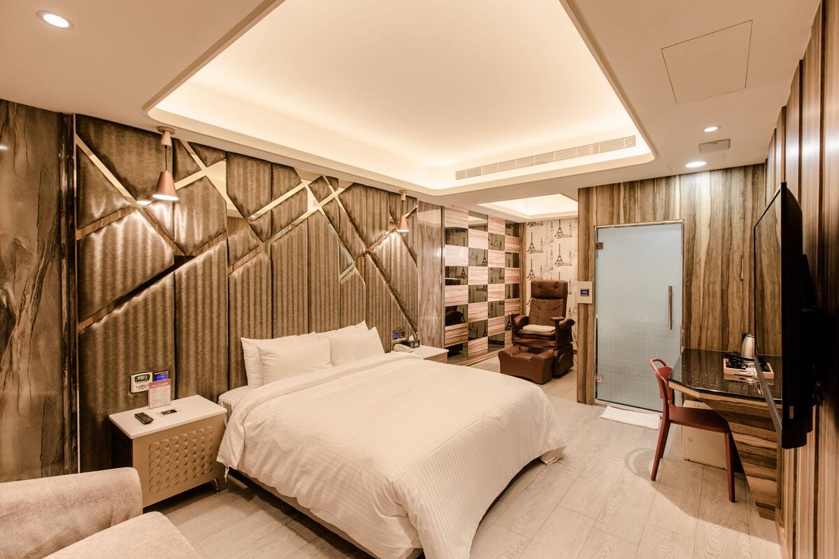A modern hotel room with a large bed, a desk with a chair, a television, and a wardrobe.