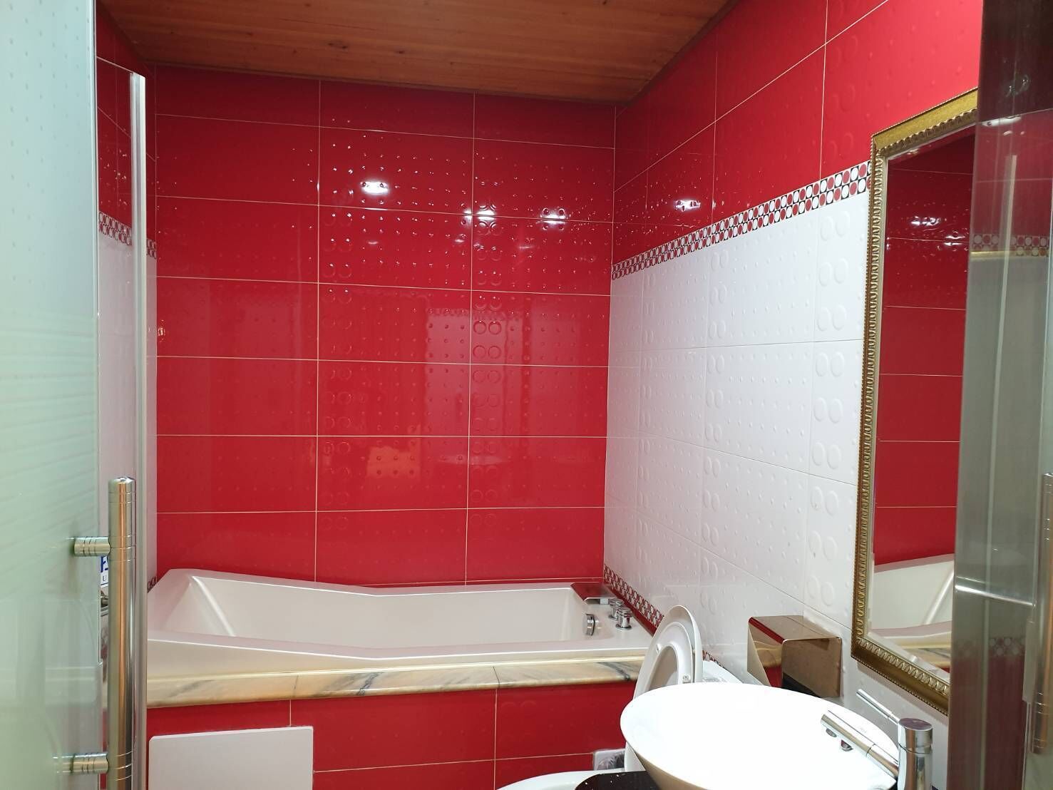 A bathroom with red tiled walls, a white bathtub, a white sink, and a mirror with a gold frame.