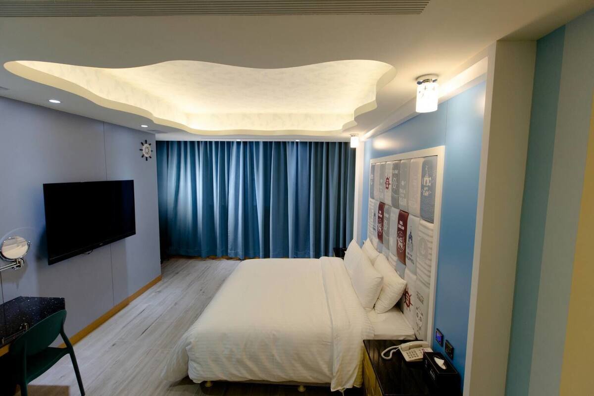 A hotel room with a bed, a flat-screen TV, a desk with a chair, a telephone, and a wall-mounted clock.