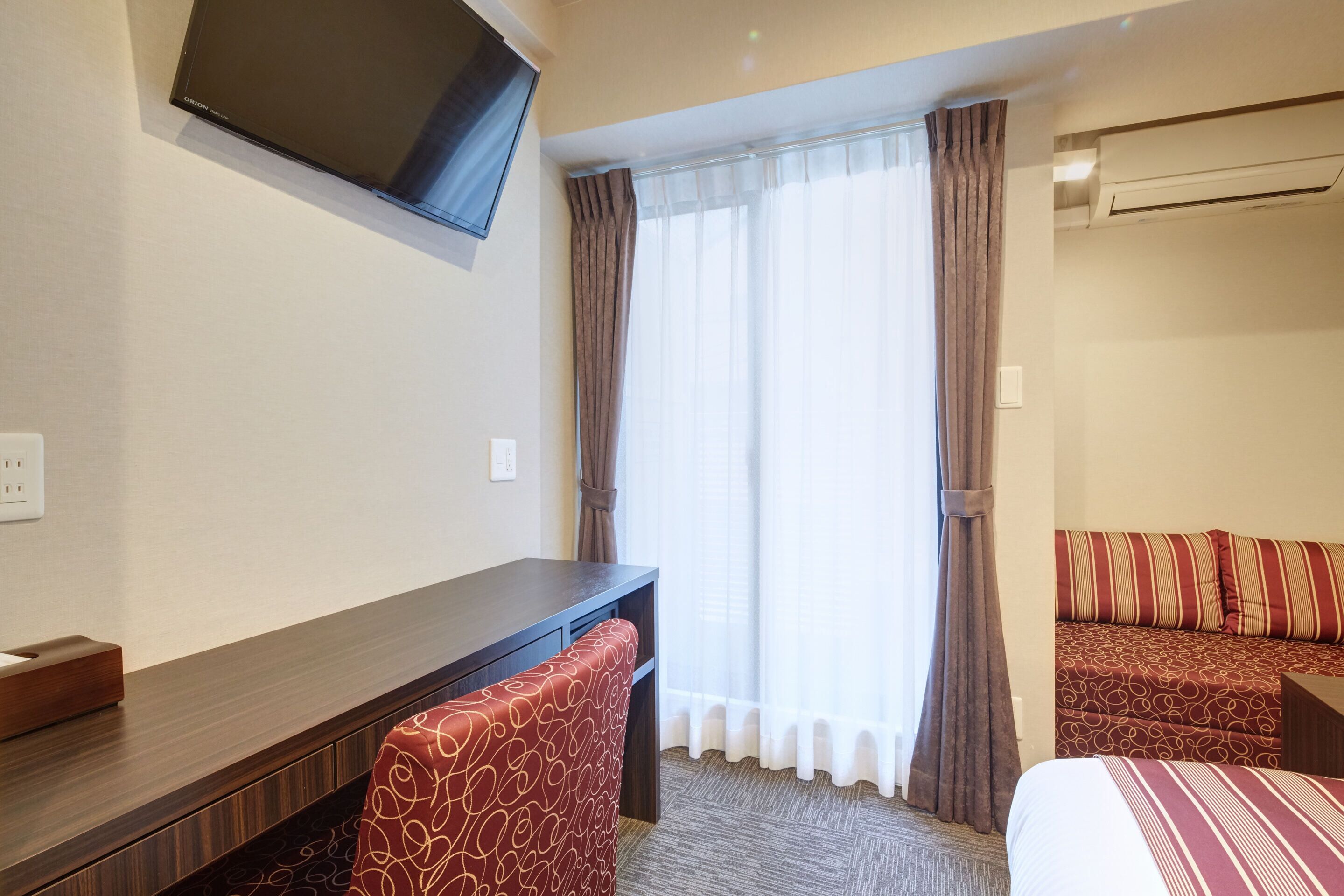 A hotel room with a bed, a desk, a chair, a TV mounted on the wall, and a window with curtains.
