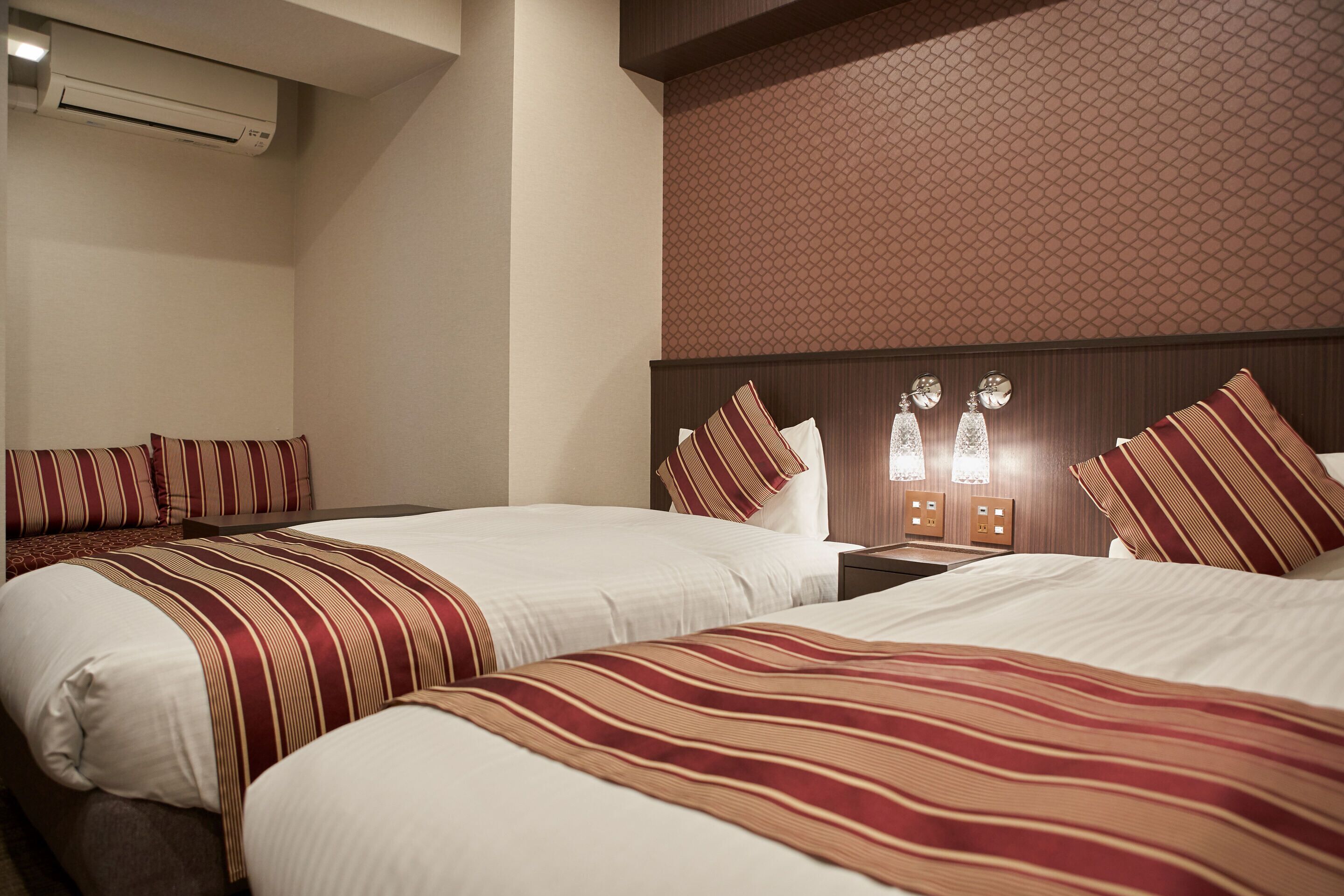 A hotel room with two beds, each with striped bedding, a wooden headboard, and a wall-mounted air conditioner.