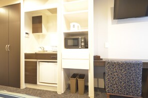 Habitación de hotel compacta con kitchenette, microondas, refrigerador pequeño, basurero y un sillón estampado.