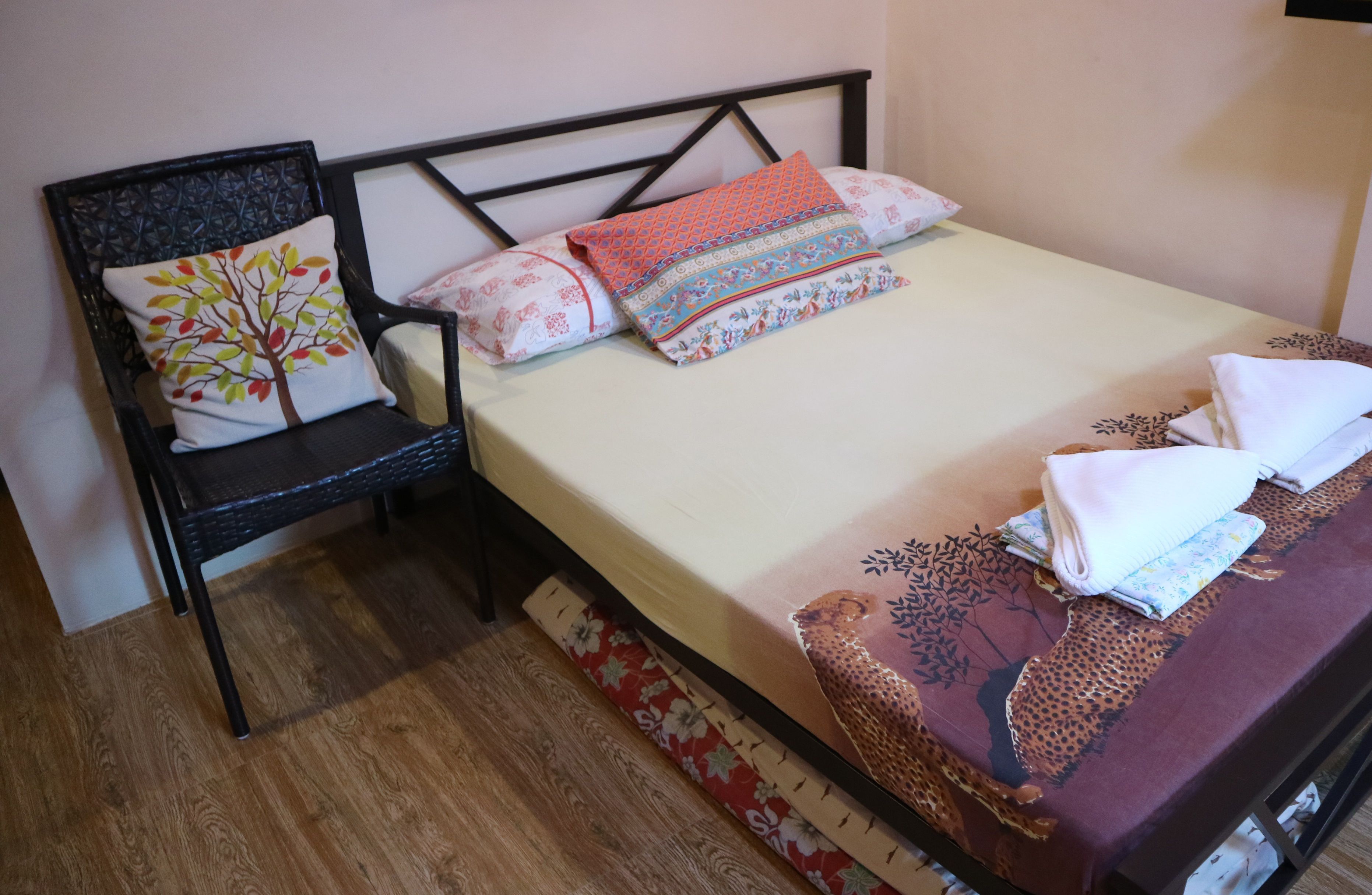 A neatly made bed with patterned bedding, a chair with a decorative pillow, and a wooden floor.