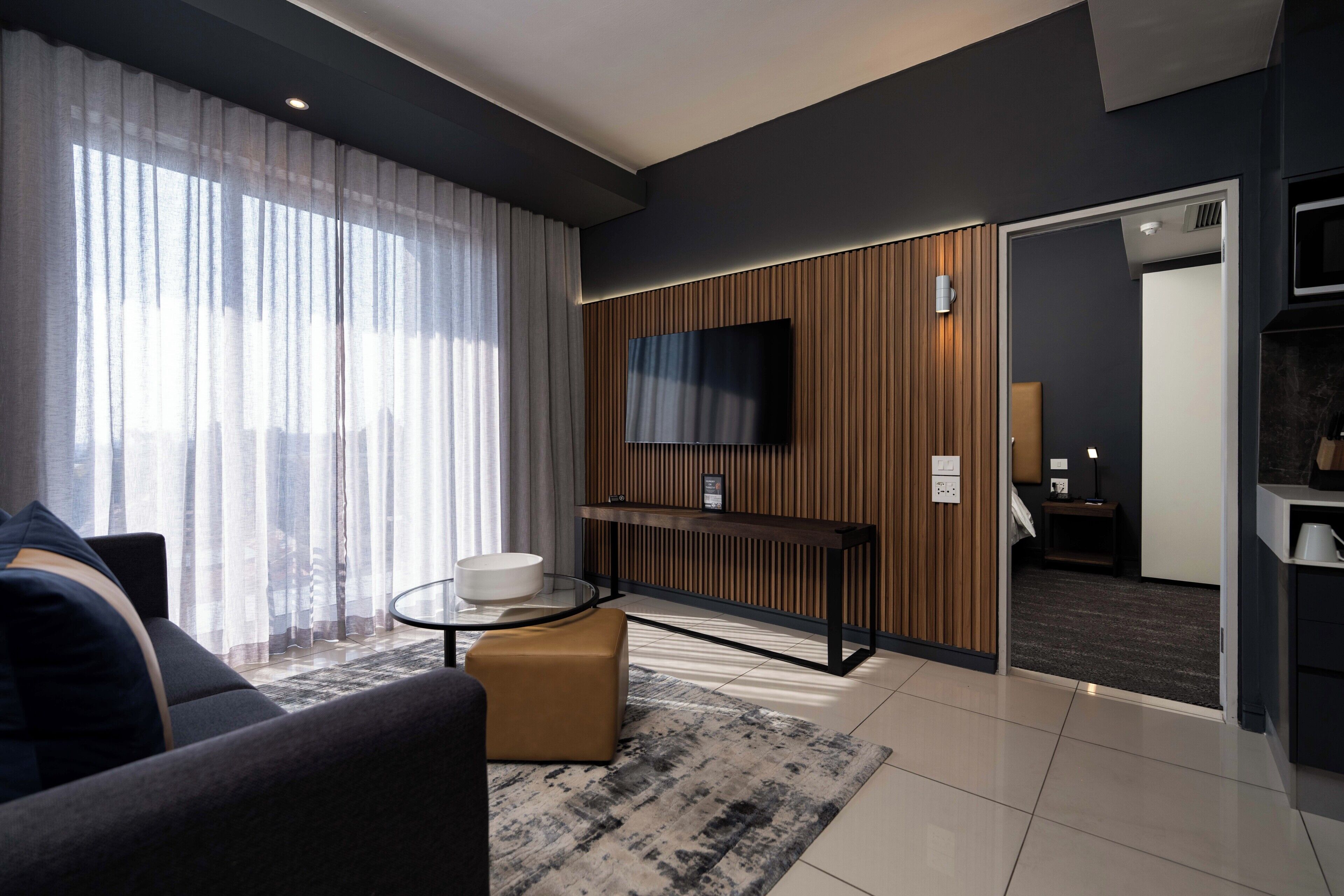 A modern living room with a sofa, a glass coffee table, a TV mounted on a wooden panel wall, and a door leading to another room.
