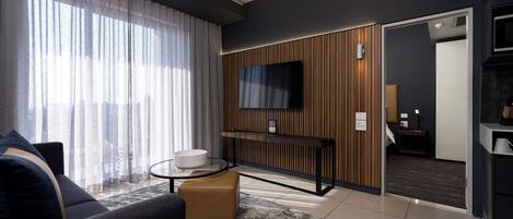 A modern living room with a sofa, a glass coffee table, a TV mounted on a wooden panel wall, and a door leading to another room.