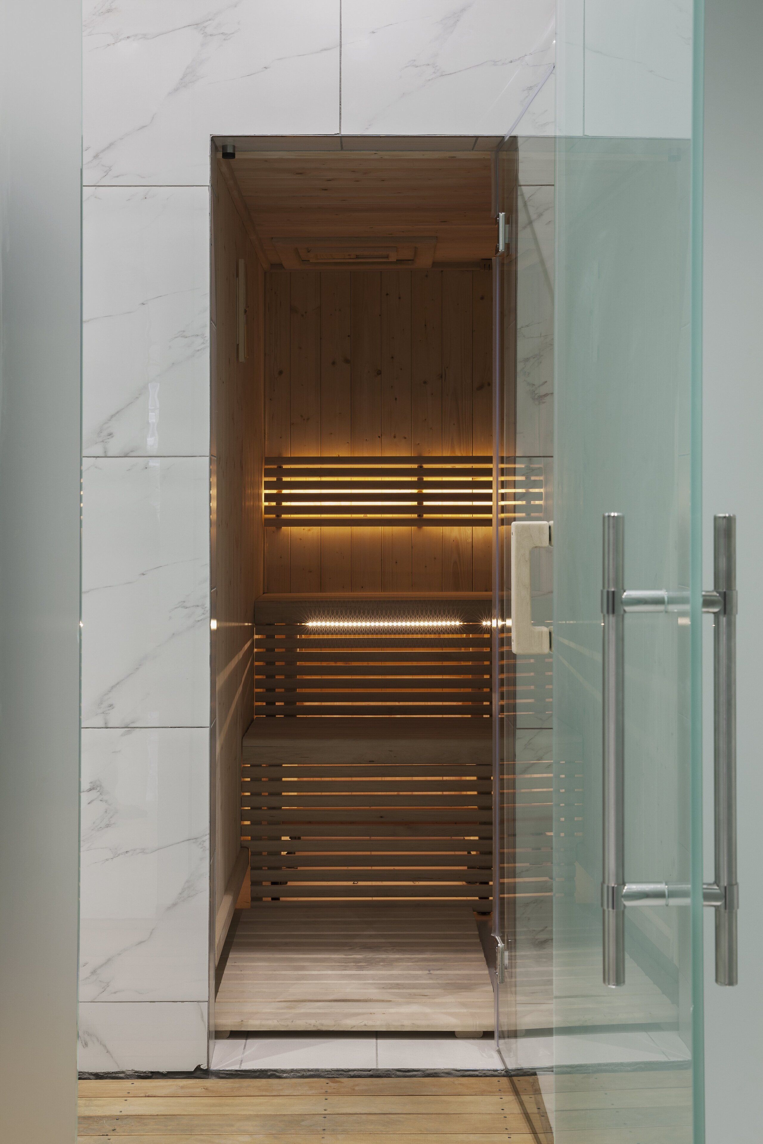 A sauna with wooden slats and a glass door, set against a marble wall.