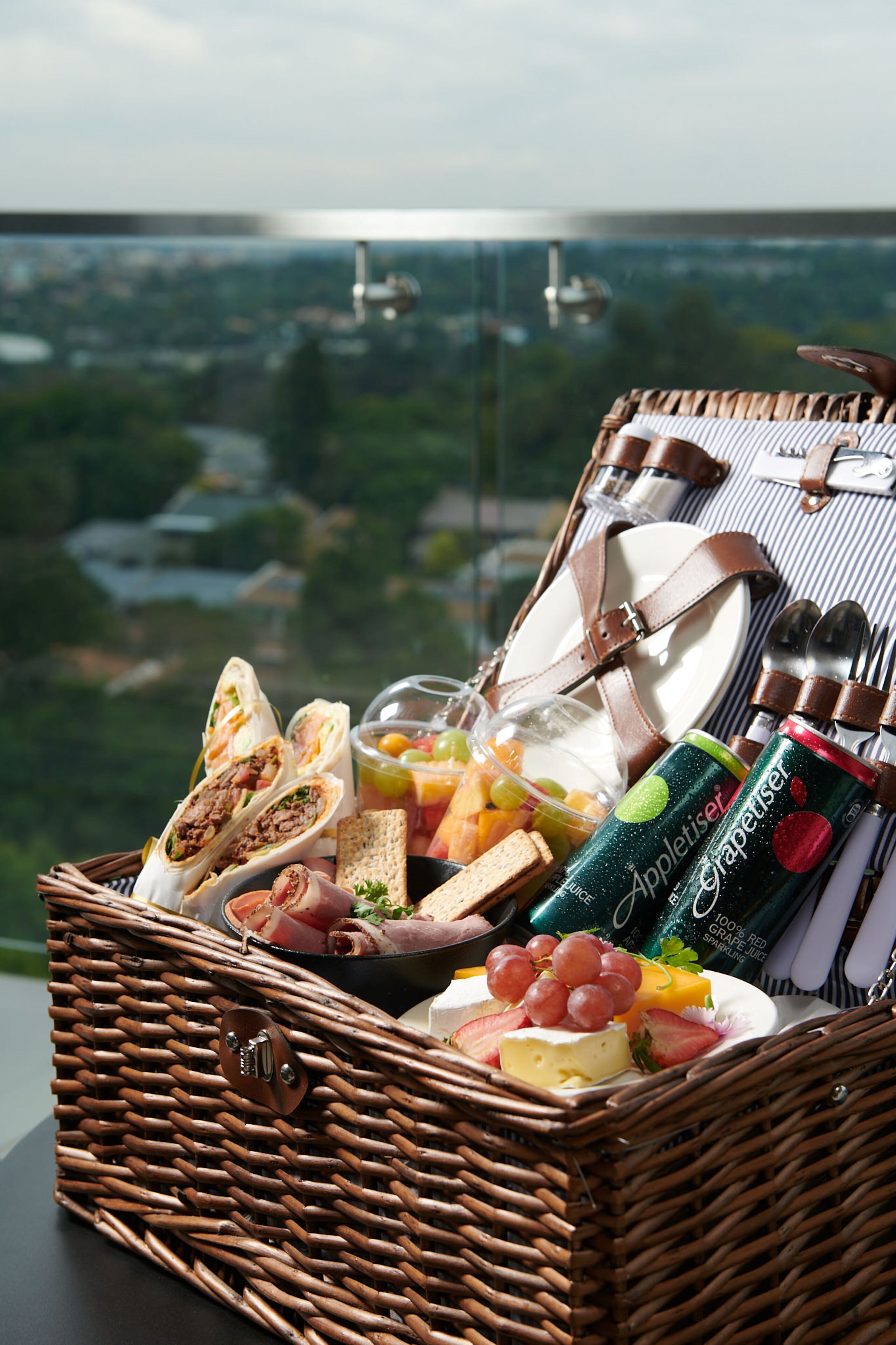 A wicker picnic basket filled with food and drinks, placed on a table with a scenic view in the background.