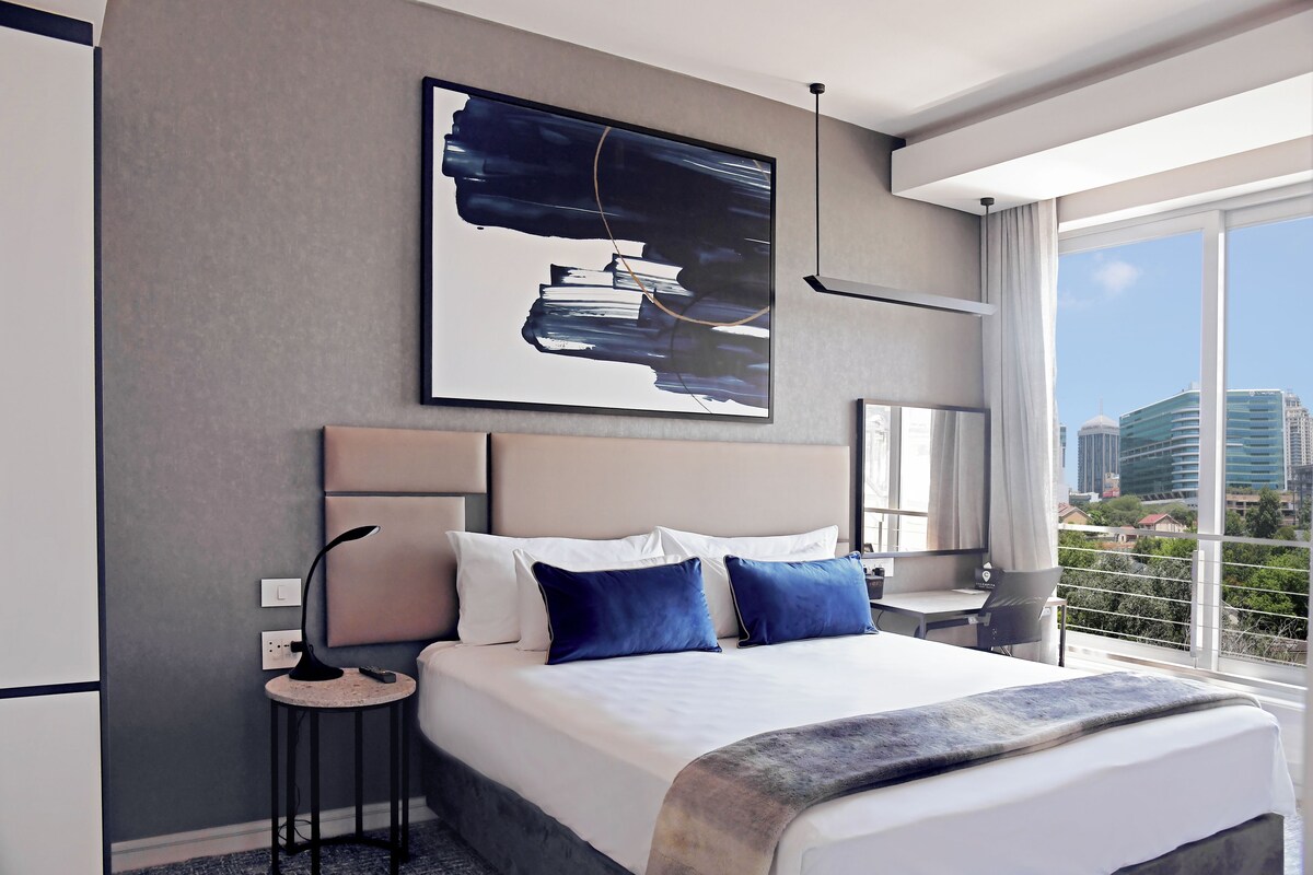 Een hotelkamer met een groot bed, een nachtkastje, een bureau, een raam met uitzicht op de stad, en een schilderij aan de muur.