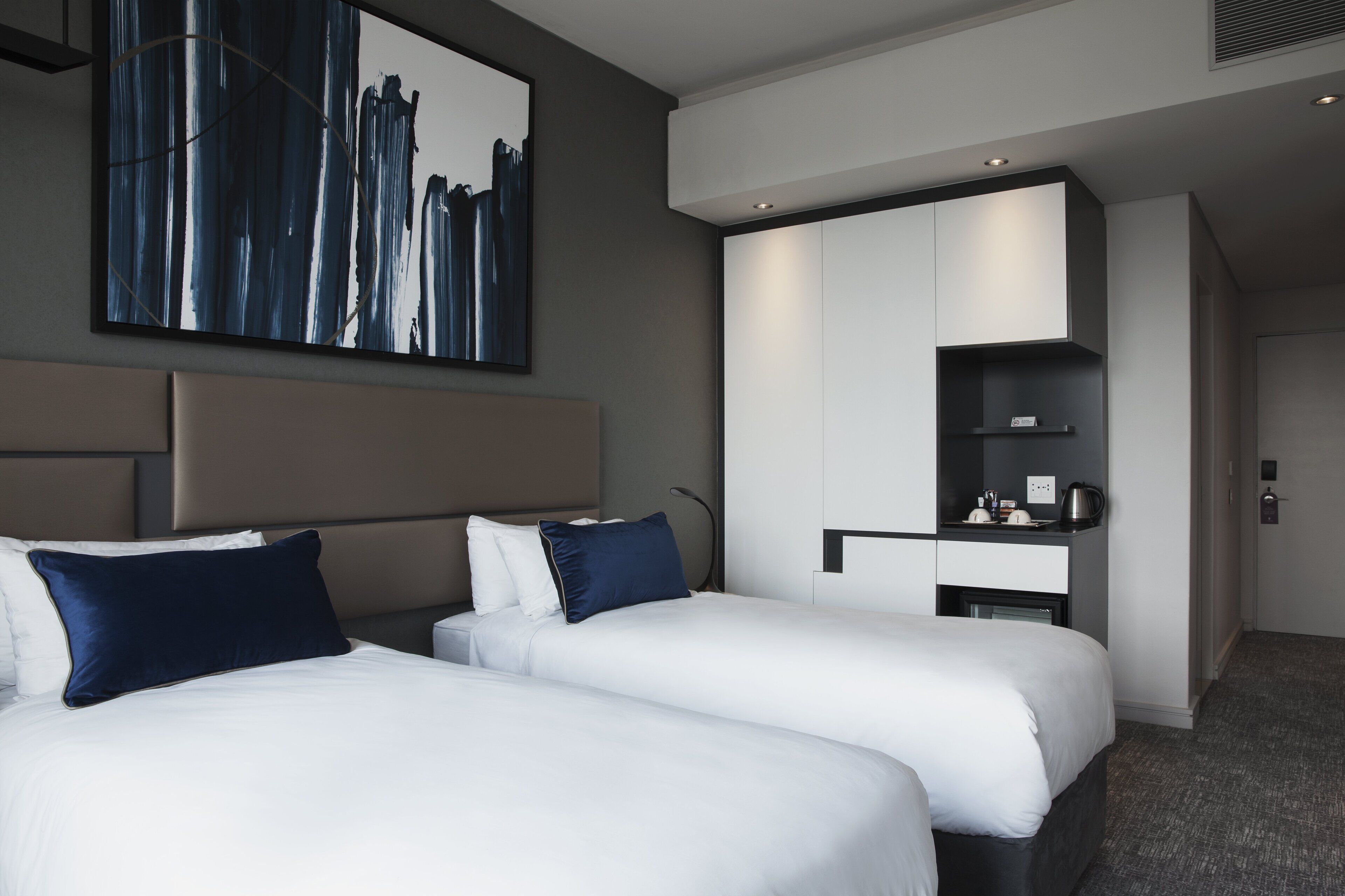 Habitación de hotel con dos camas, un cuadro abstracto grande, un armario moderno y una kitchenette pequeña.
