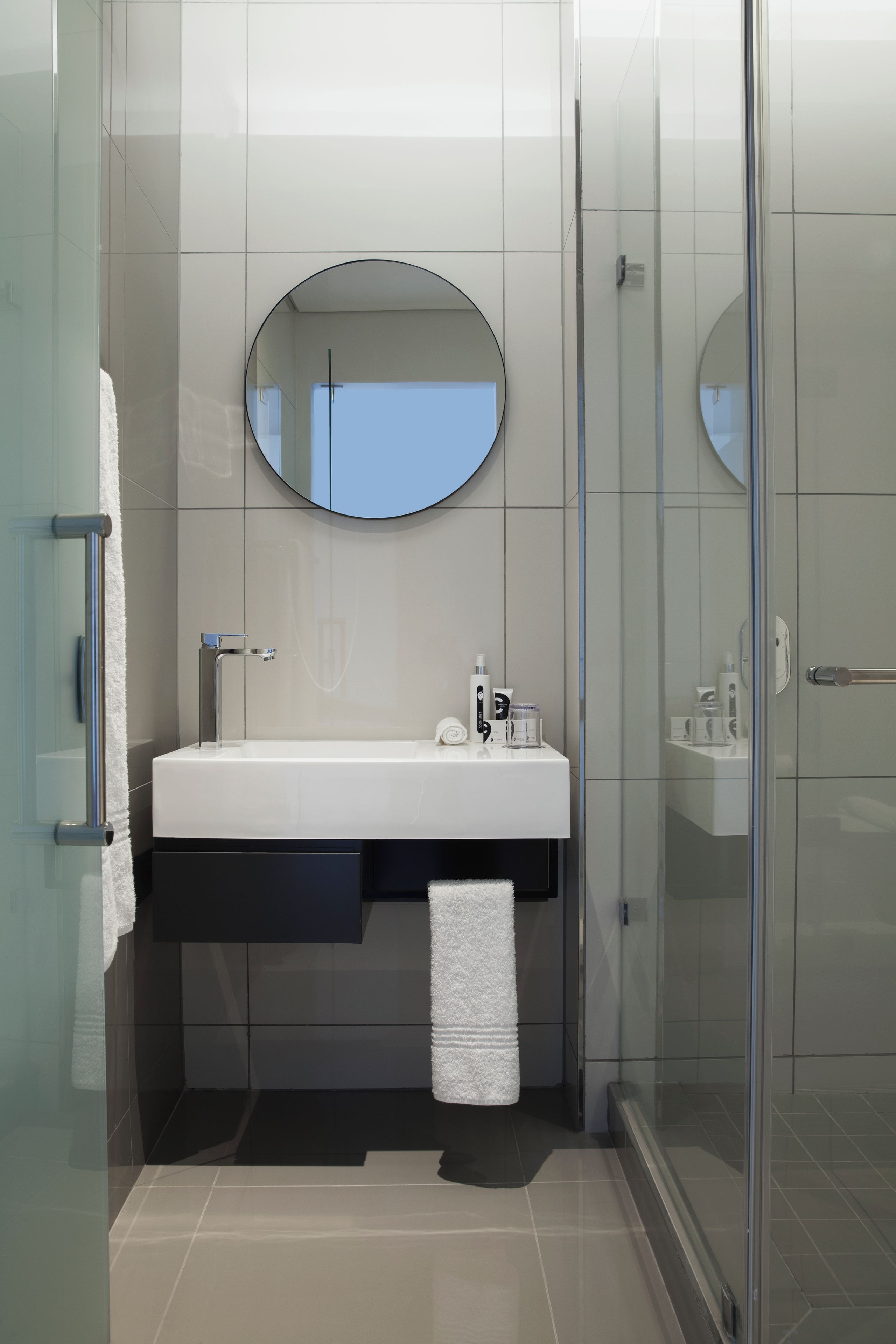 A modern bathroom with a glass shower enclosure, a white sink with a round mirror above it, and a towel hanging on the wall.